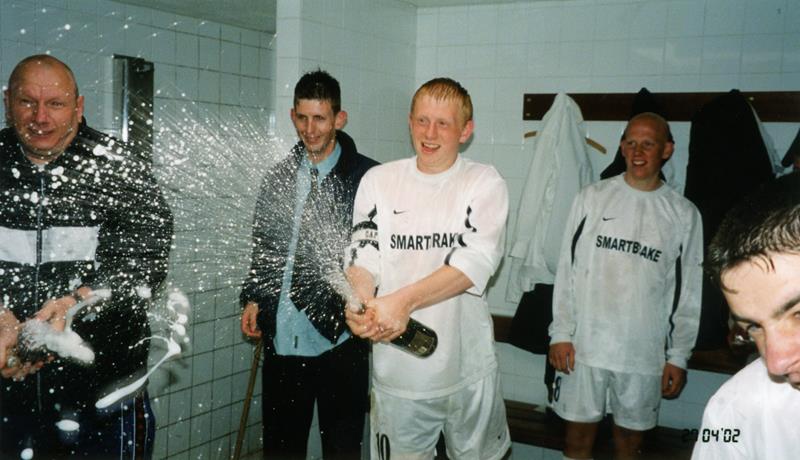 Predators celebrating a cup win in the dressing room with a player opening a bottle of champagne looked on by manager Big Paul Harris.